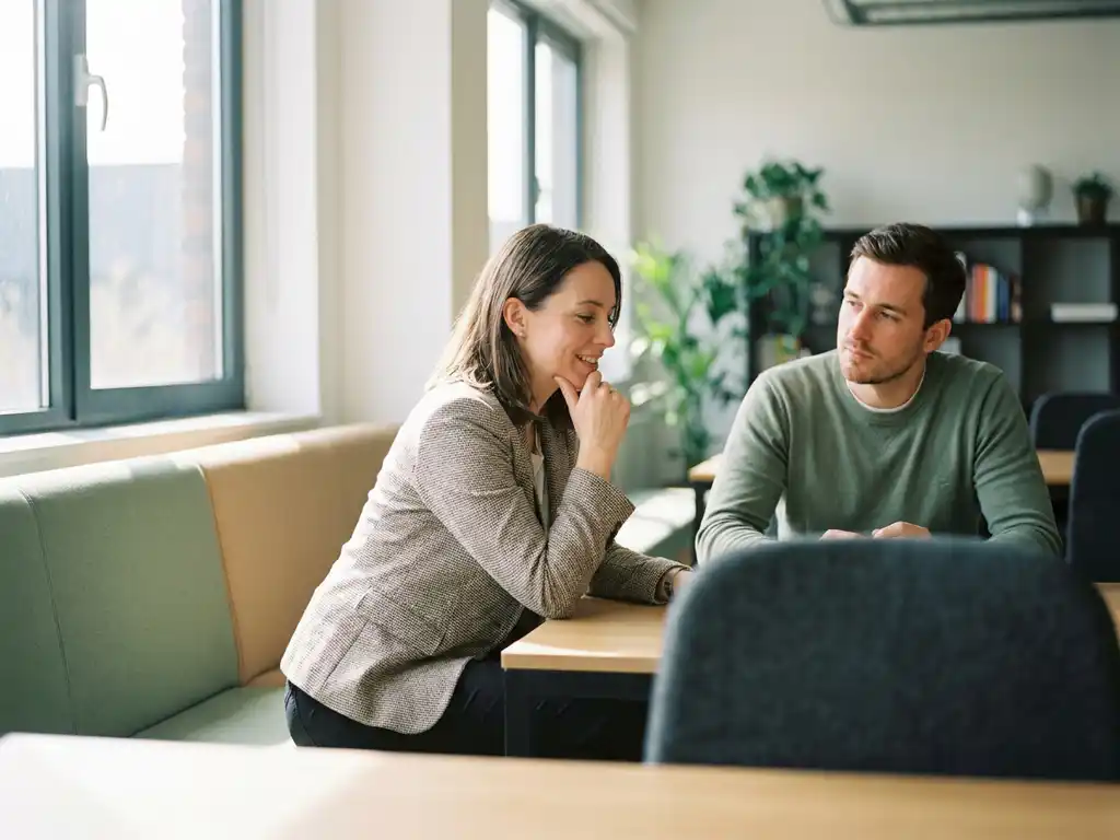 Professional leunt nieuwsgierig voorover tijdens gesprek met collega in modern kantoor met zacht natuurlijk licht.